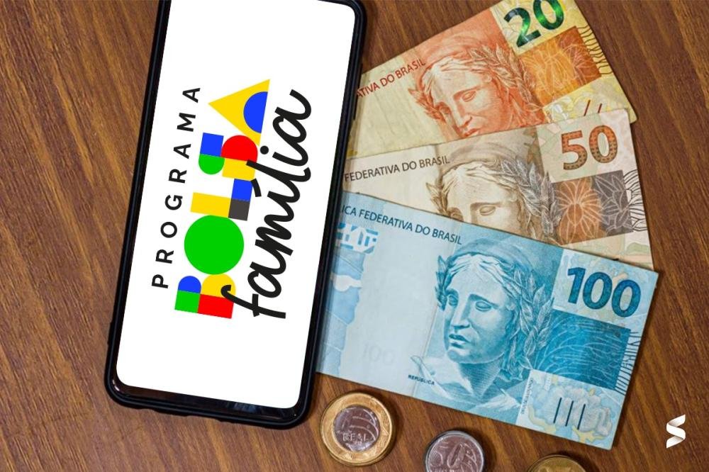 Cartão do Bolsa Família sendo segurado por uma pessoa com notas de 100 reais ao fundo