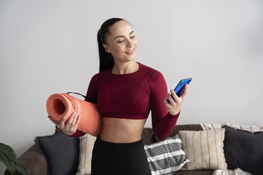 Mulher segurando tapete de yoga enquanto olha para o celular, equilibrando exercícios físicos e uso de tecnologia para emagrecimento