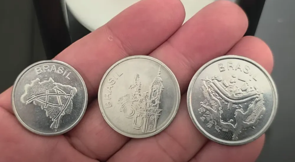 Três moedas brasileiras antigas na palma da mão, exibindo o mapa do Brasil, uma igreja barroca e o brasão nacional.