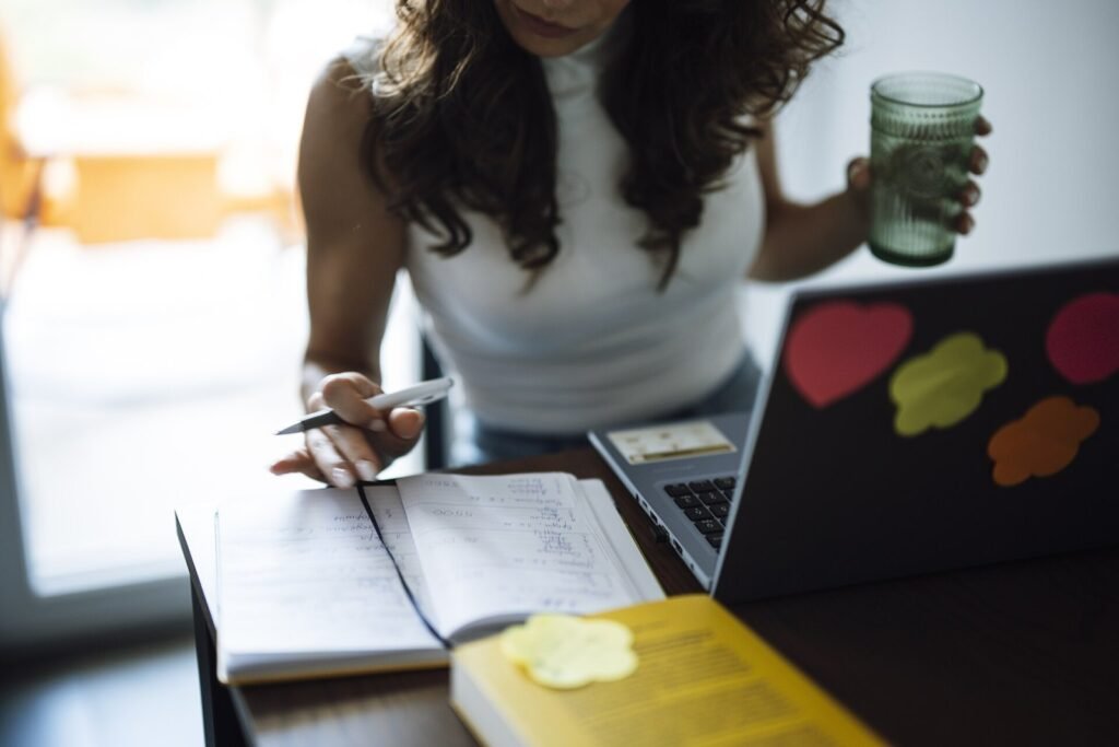 A woman sits at a desk working on a laptop while holding a green drink. She writes notes in a notebook. Sticky notes are placed on the laptop. The roo