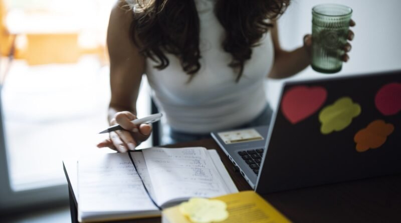 A woman sits at a desk working on a laptop while holding a green drink. She writes notes in a notebook. Sticky notes are placed on the laptop. The roo