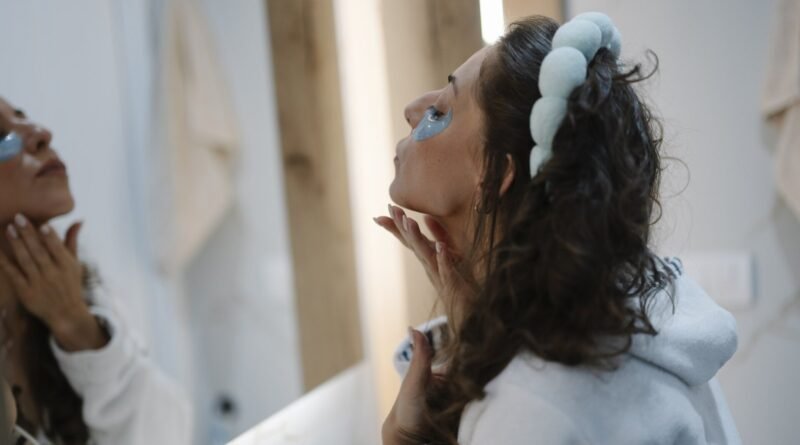 A woman is standing in front of a mirror in a bathroom. She is using a skincare product on her face while dressed in comfortable clothes. The setting